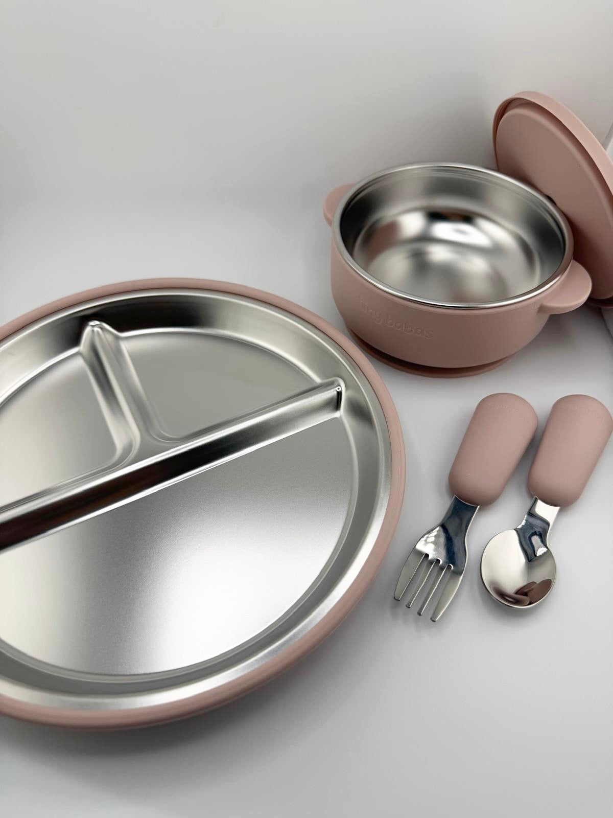 A baby feeding set with a divided stainless steel plate, a small stainless steel bowl with a pink lid, and matching pink utensils including a fork and spoon, arranged on a light gray background.
