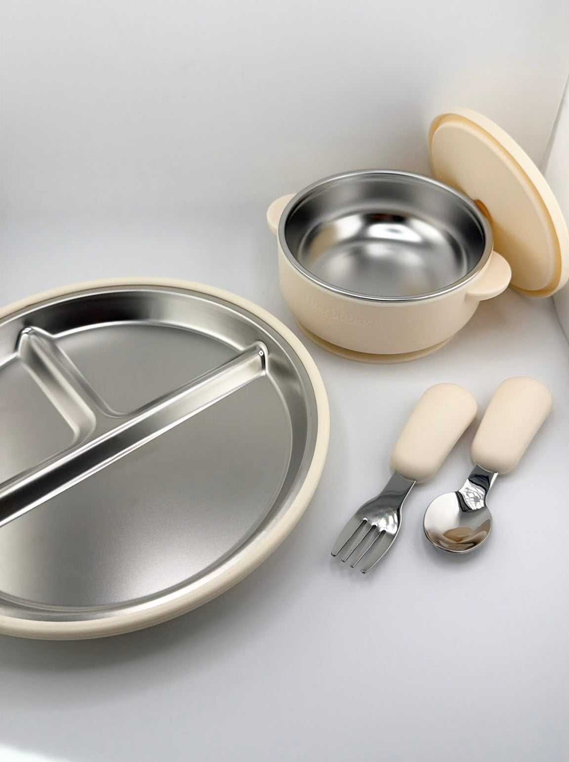 A baby feeding set including a divided stainless steel plate, a small stainless steel bowl with a cream-colored lid, and matching utensils with cream-colored handles, arranged on a light gray surface.
