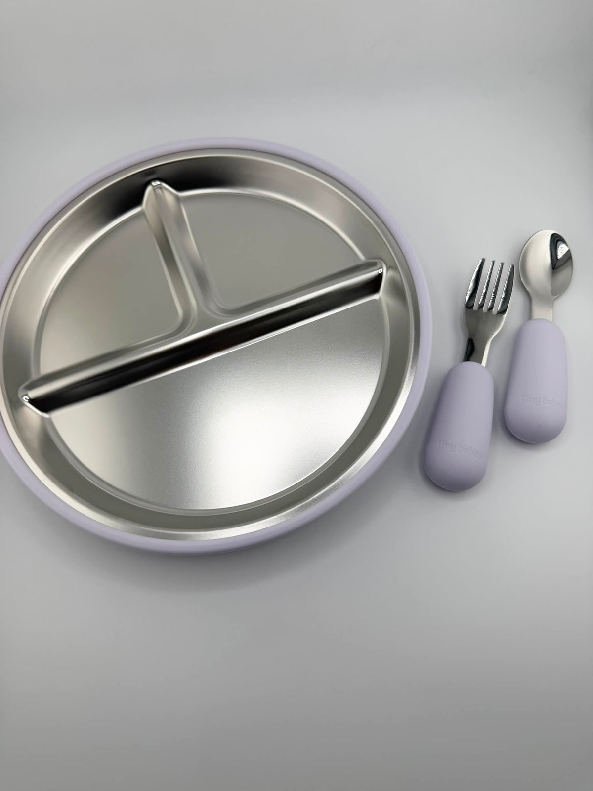 A stainless steel divided baby plate with a light purple rim, shown with matching fork and spoon that have light purple handles on a neutral gray background.