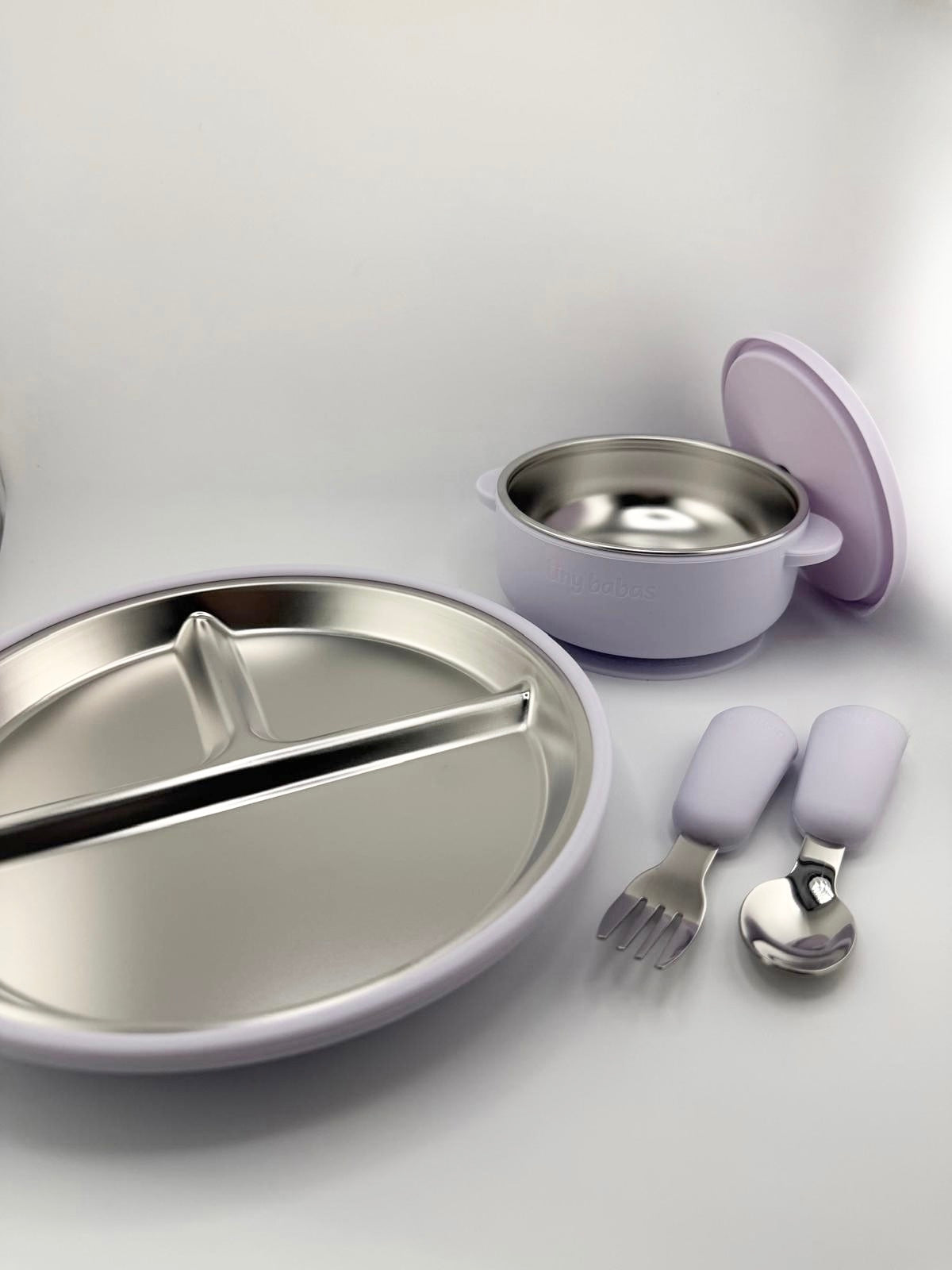 A children's dinnerware set in lavender and stainless steel, including a divided plate, a small bowl with lid, and a fork and spoon with lavender handles, arranged on a light gray background.