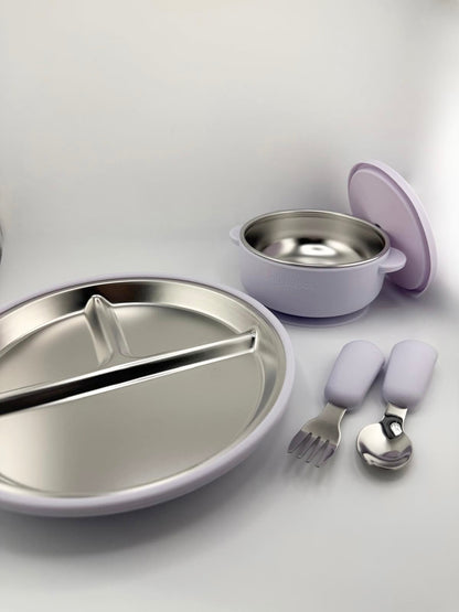 A children's dinnerware set in lavender and stainless steel, including a divided plate, a small bowl with lid, and a fork and spoon with lavender handles, arranged on a light gray background.