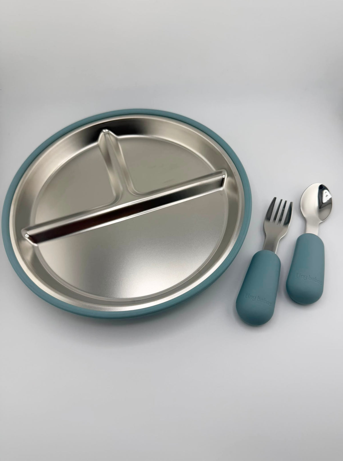 A stainless steel divided baby plate with a teal silicone rim, shown with a matching fork and spoon that have teal handles on a light gray background.
