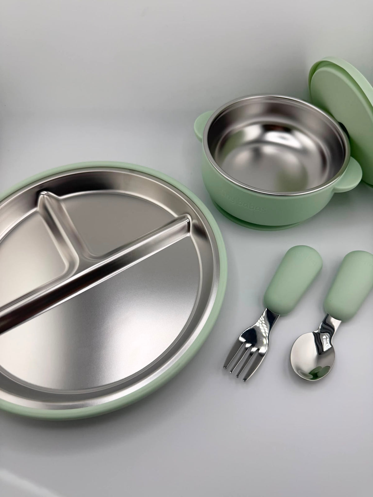 A children's stainless steel dinner set with mint green handles, including a divided plate, a small bowl with lid, and a fork and spoon, arranged on a light gray surface.