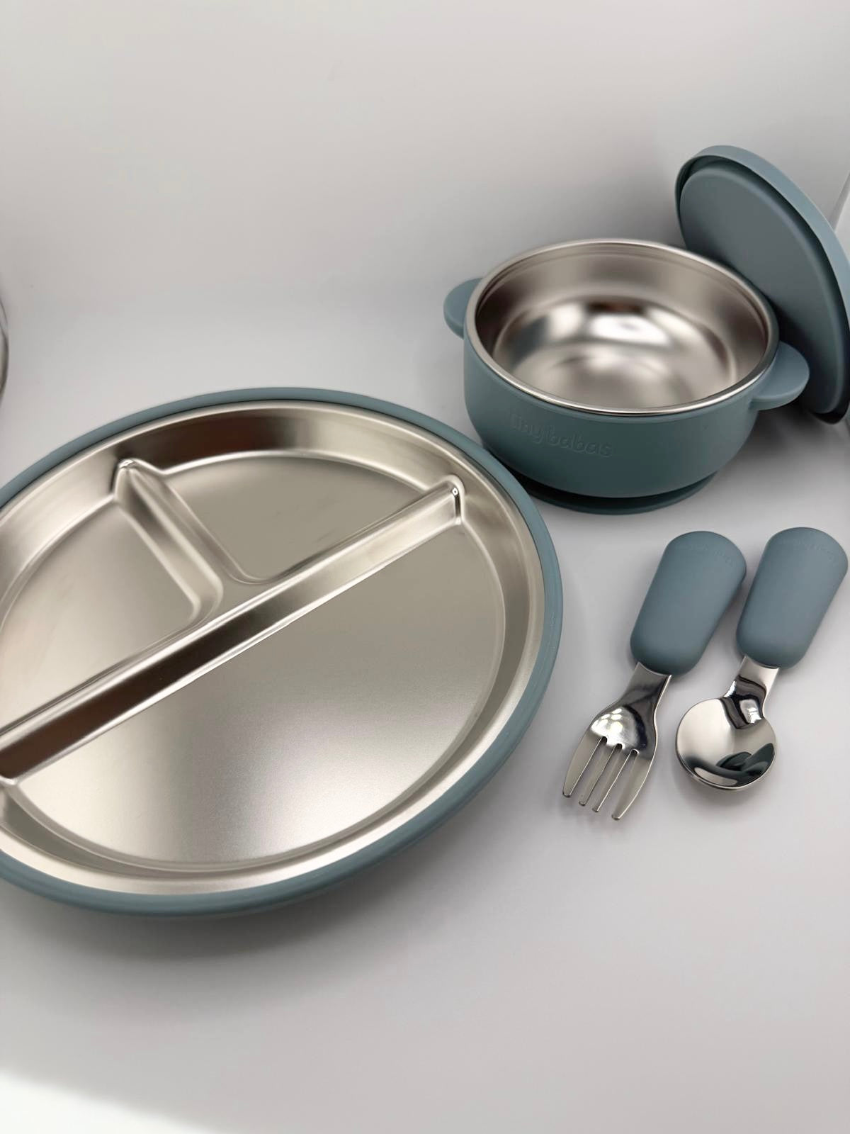 A children's stainless steel dinner set with a divided plate, a small bowl with lid, and matching utensils with blue-gray handles, arranged on a light gray surface.
