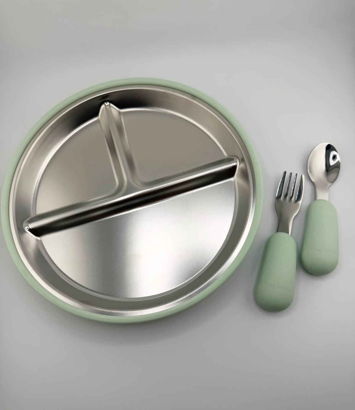 A stainless steel divided baby plate with a mint green silicone rim, shown with matching silicone-handled utensils on a light gray background.