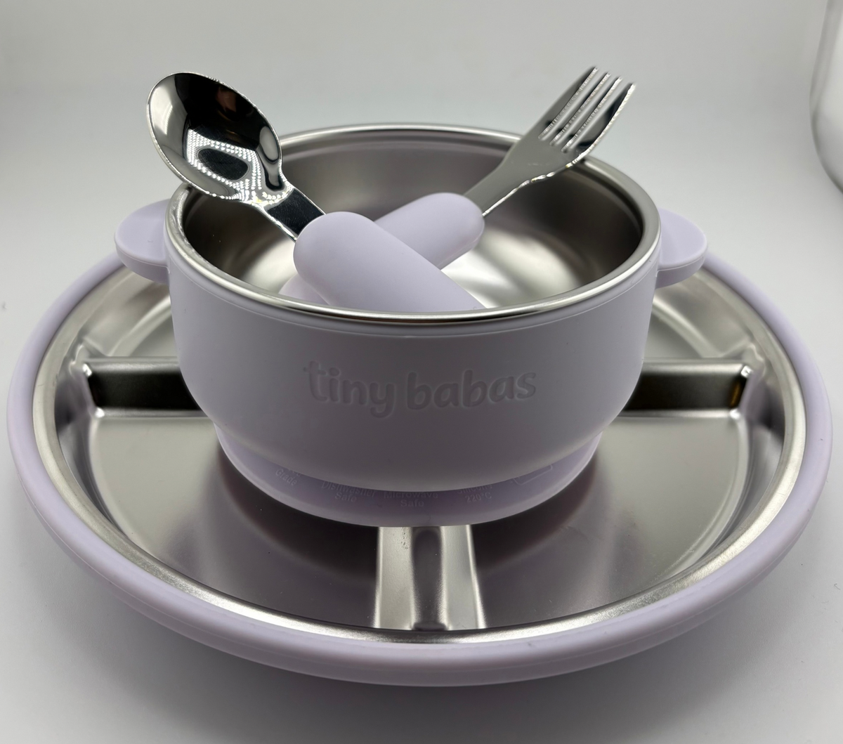 A baby feeding set by tiny babas, featuring a lavender silicone suction bowl with a stainless steel interior, accompanied by a matching silicone spoon and fork with lavender handles, all resting on a stainless steel plate.