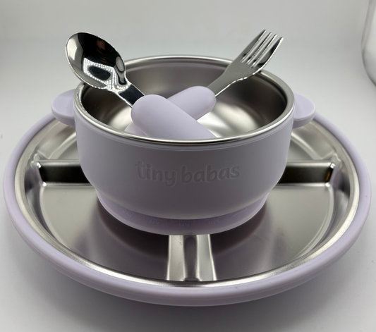 A baby feeding set by tiny babas, featuring a lavender silicone suction bowl with a stainless steel interior, accompanied by a matching silicone spoon and fork with lavender handles, all resting on a stainless steel plate.